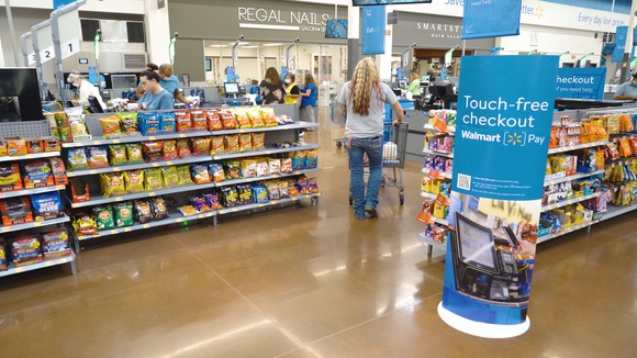 A customer pushes a cart through the aisles of a walmart store with a sign promoting touch-free checkout.