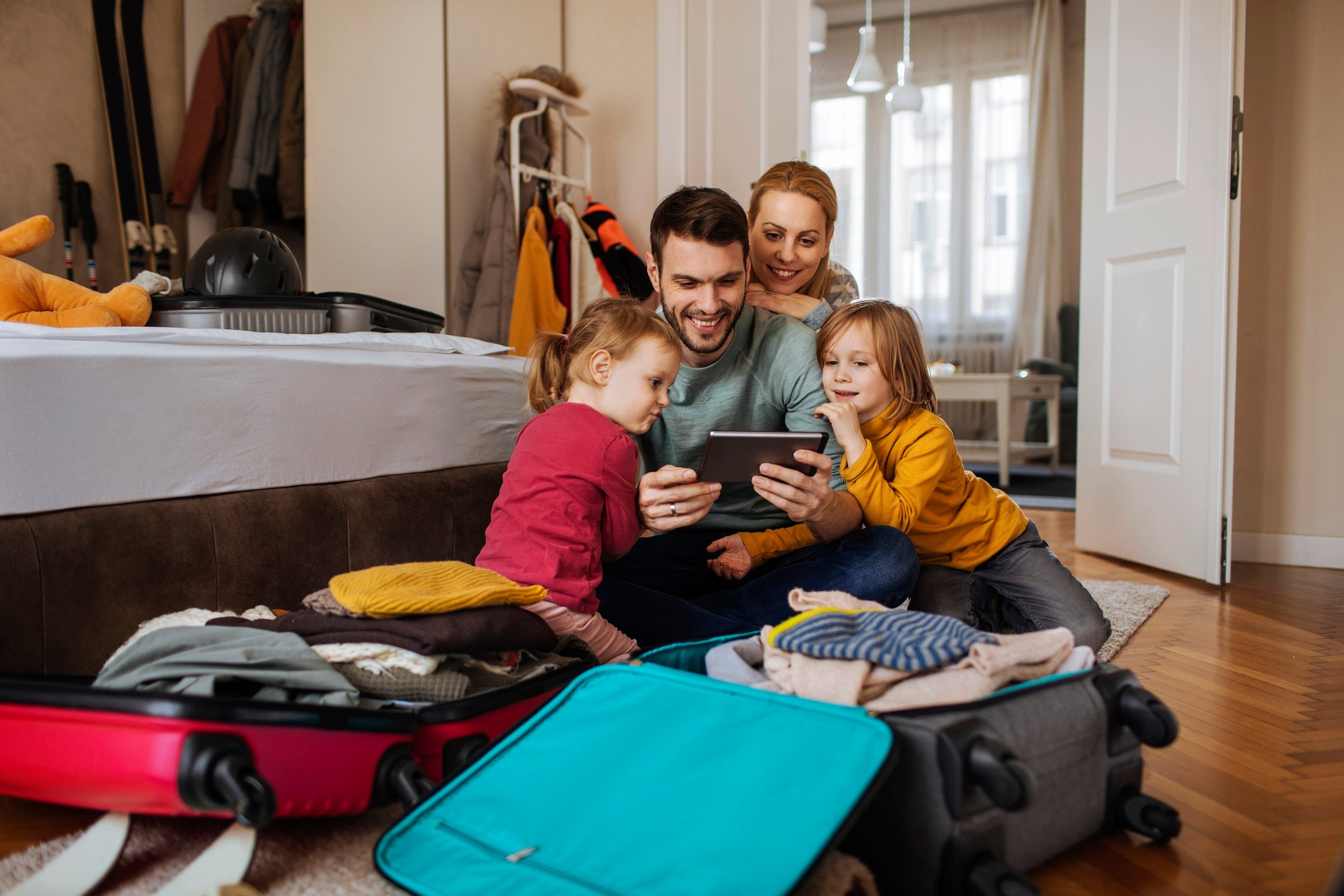 Husband, wife, and two young daughters gathered around a tablet after opening suitcases at their vacation rental.