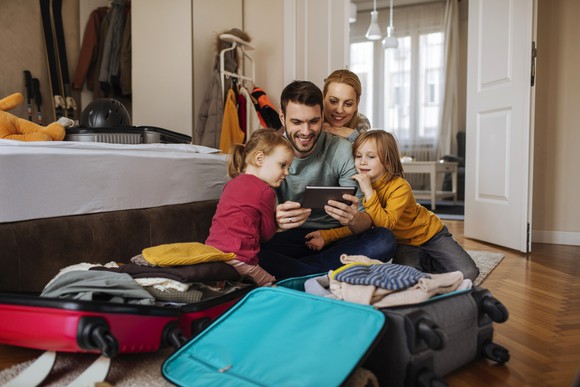 Husband, wife, and two young daughters gathered around a tablet after opening suitcases at their vacation rental.