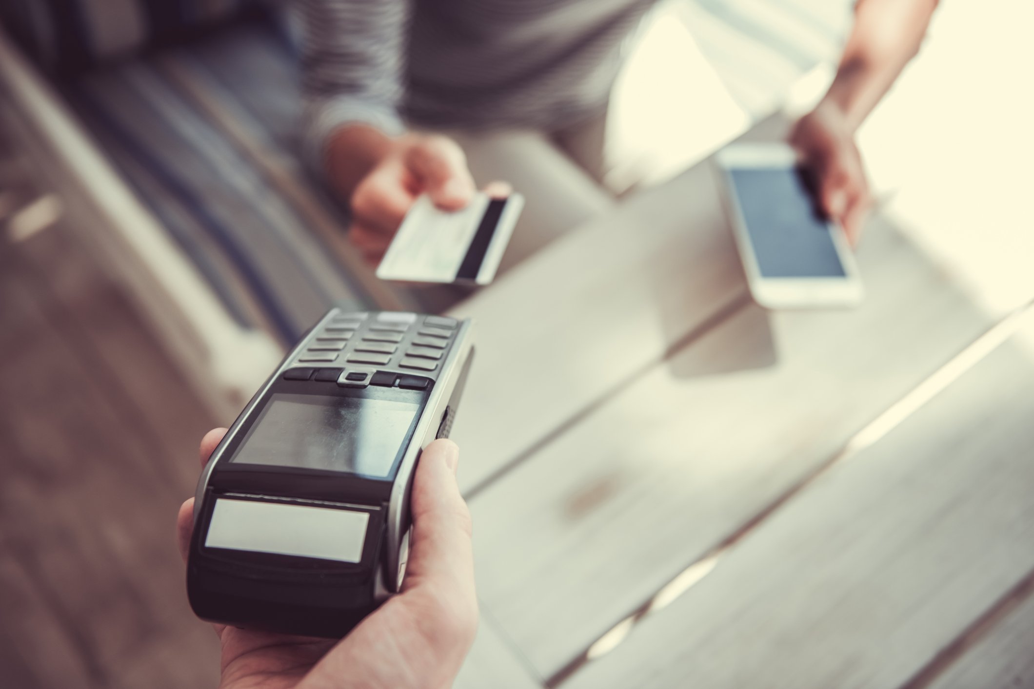 Person with a credit card and a smartphone making a payment at a point-of-sale terminal.