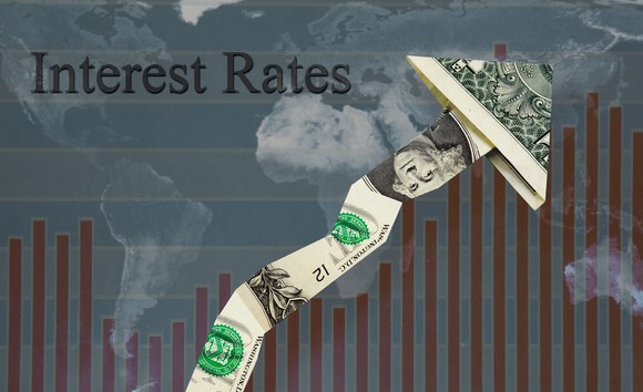 A rising line with an arrow made out of folded one dollar bills, with the word interest rates in the background.