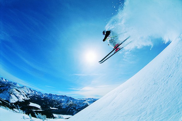 A man skiing down a mountain under a blue sunny sky with the sun centered, in a cloud of powdery snow.