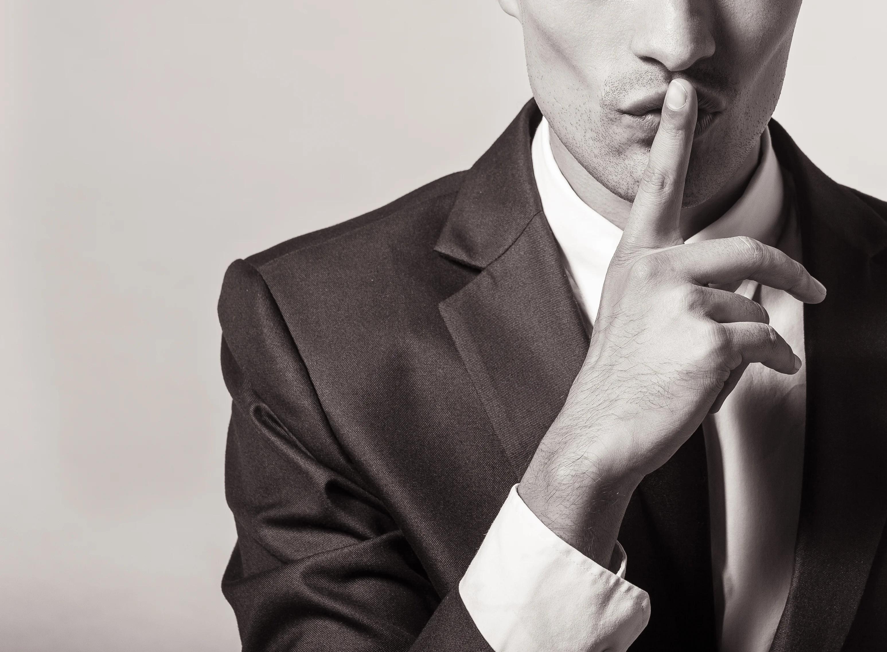 Person in suit with finger over his lips signaling 'Keep quiet.'