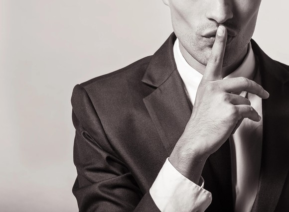 Person in suit with finger over his lips signaling 'Keep quiet.'