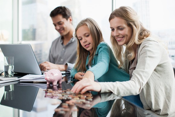 A mother and her daughter count coins in front of a piggy bank while a father consults a computer in the background.