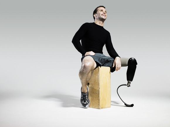 An athlete with a prosthetic leg smiles as he sits on a block of wood.