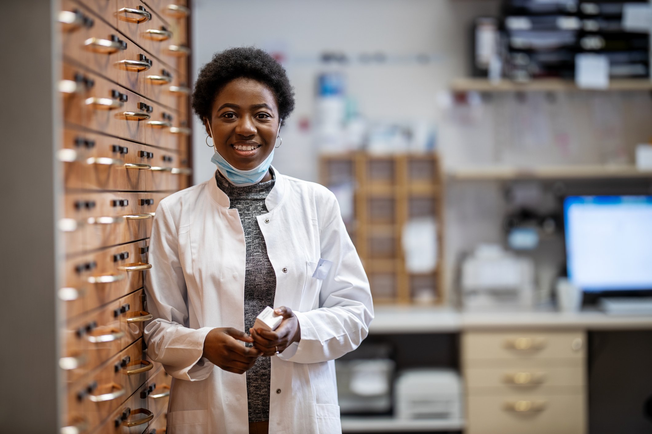 Female pharmacist in a white coat holds a medicine bottle while leaning against a wall of medicine drawers and files.