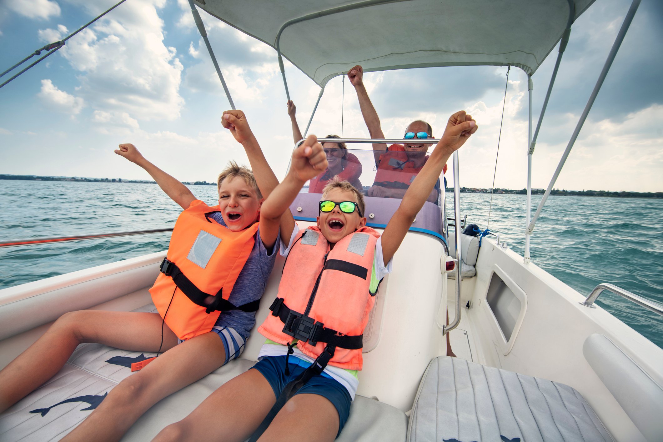 A family on a boat with kids sitting up front wearing life vests and their hands in the air.