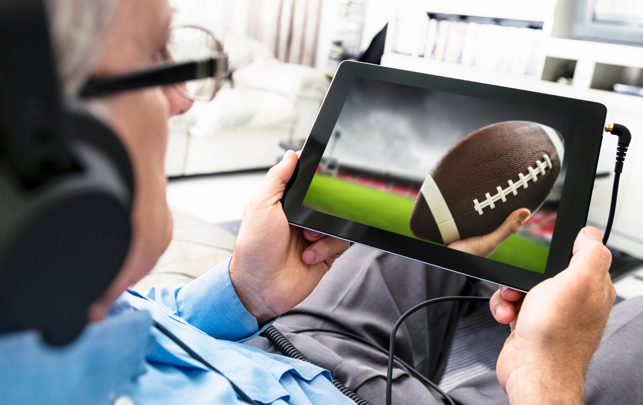 A man using a tablet to watch a digital sportscast, with an image of a football on the screen.