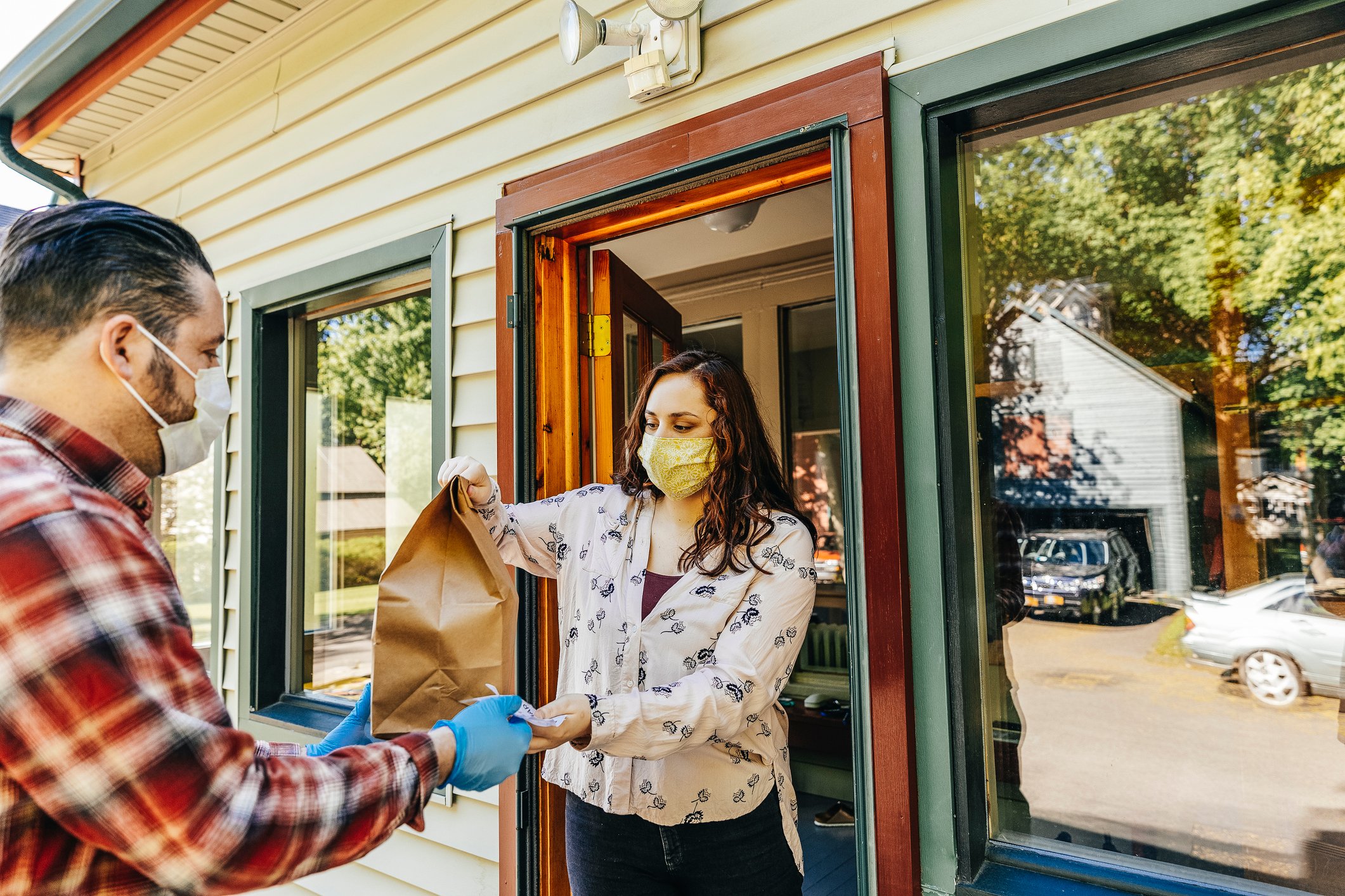 A masked and gloved young man handing a masked woman a food delivery bag.