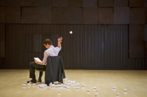 A businessman sits in an empty auditorium throwing crumpled papers behind his back.