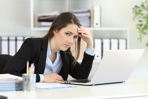 A young female office worker at her laptop, glancing at the camera with a frown.