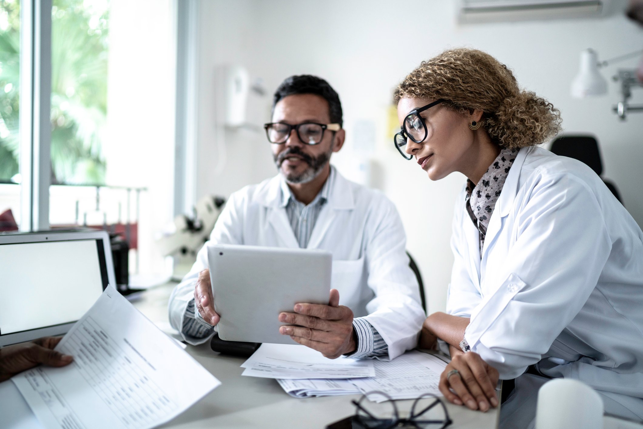 Male and female doctors  discuss around paperwork and a tablet at an office desk.