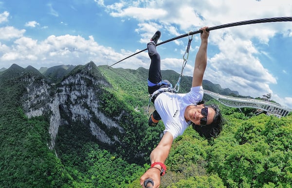 A daredevil takes a selfie while dangling from a steel cable above a wide, deep mountain gorge.
