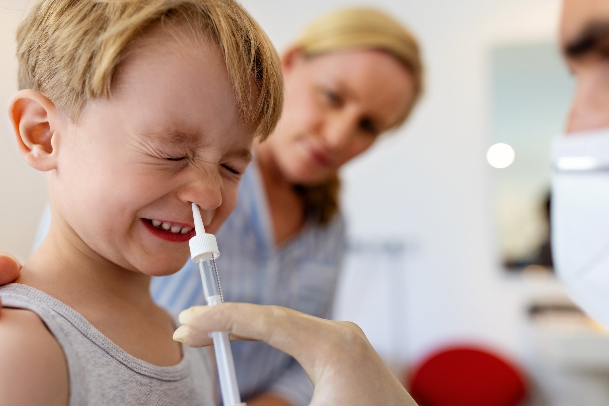 A child squinting and preparing to get a flu vaccine nasal spray with his mother looking on in the background.