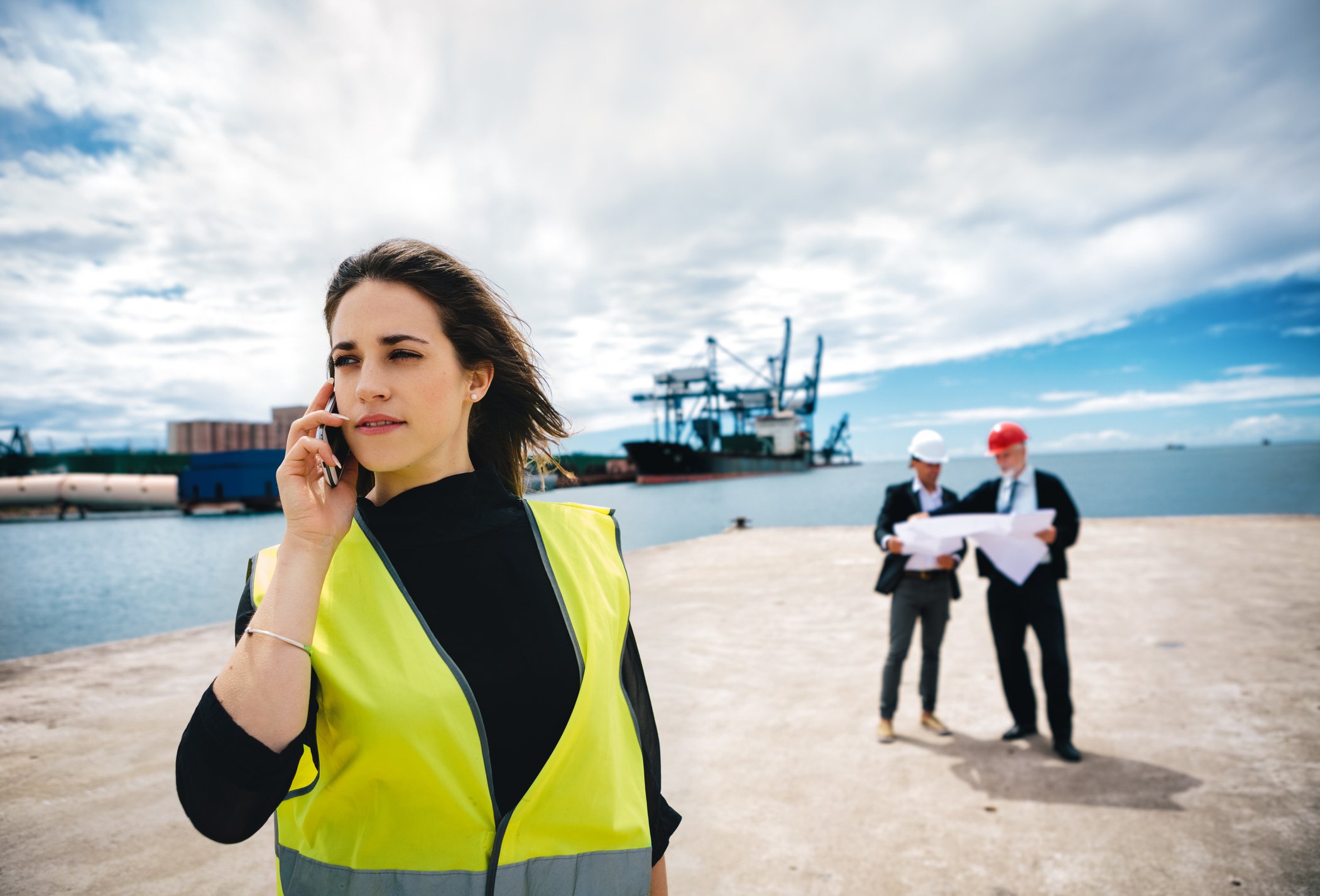 Woman clad in yellow safety vest using a mobile phone at a dock.