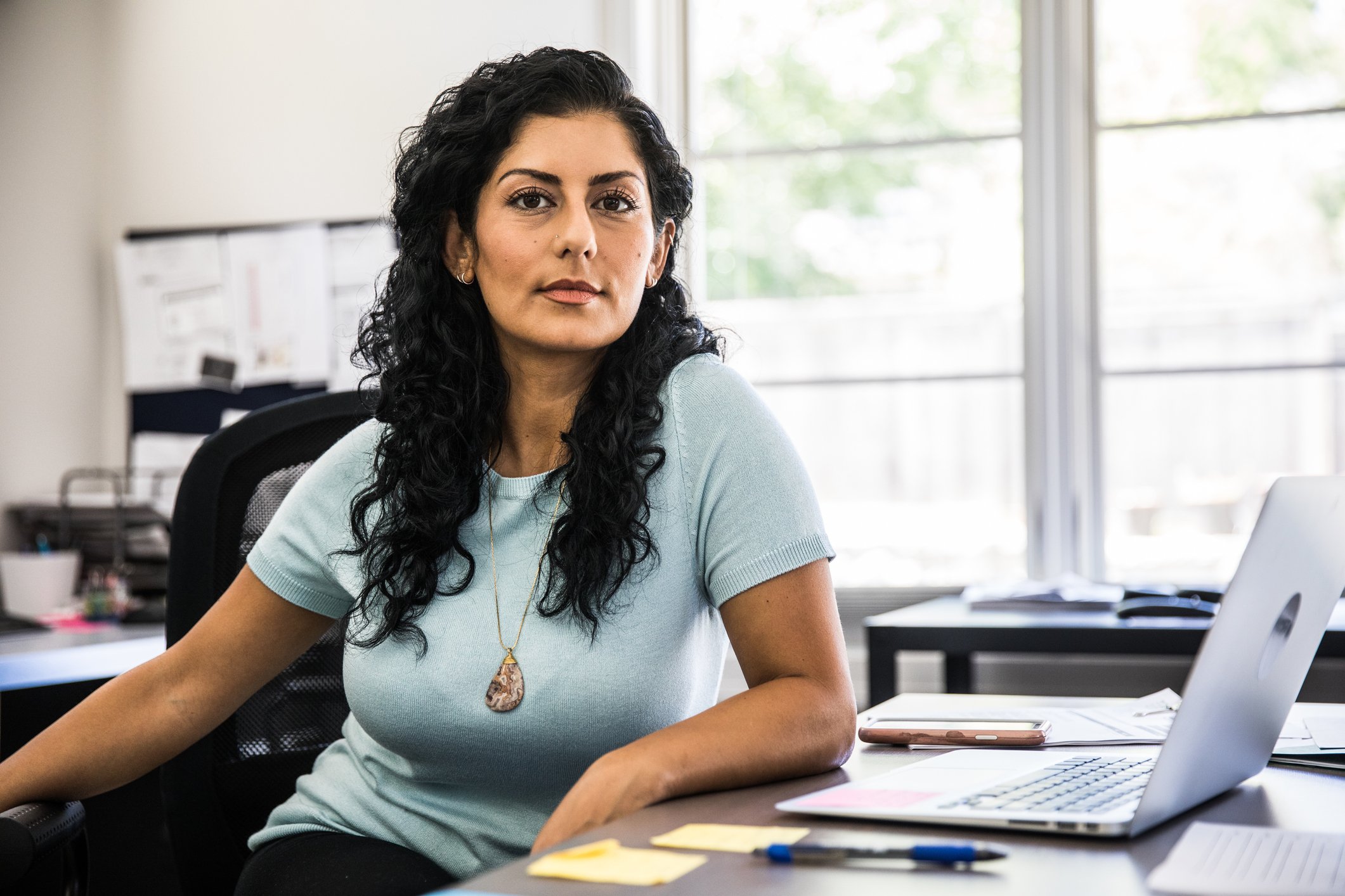 Woman sits at a business office desk in front of a sticky pad of notes, cell phone, and laptop.