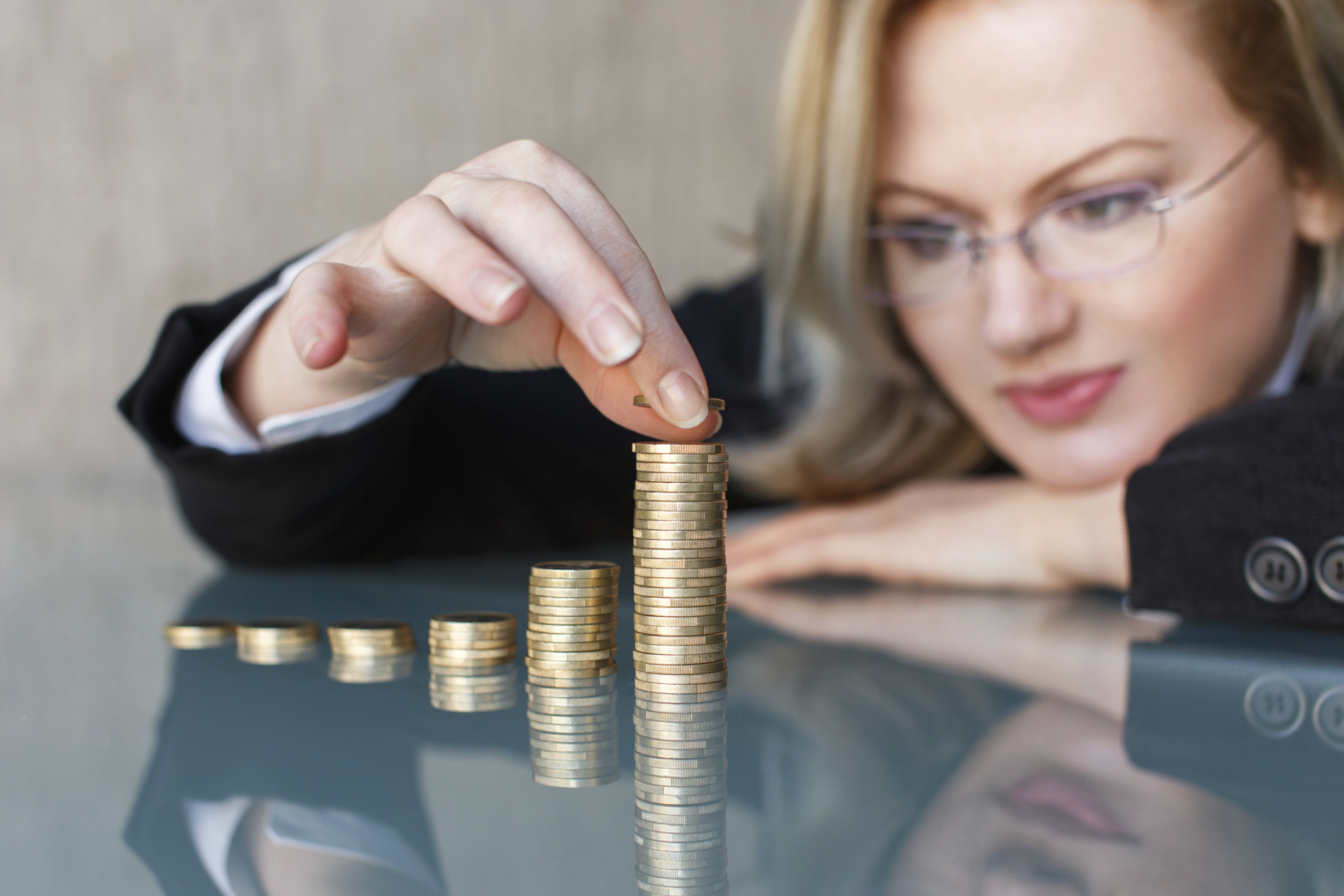 A young businesswoman stacks coins in rising piles on a glass tabletop.