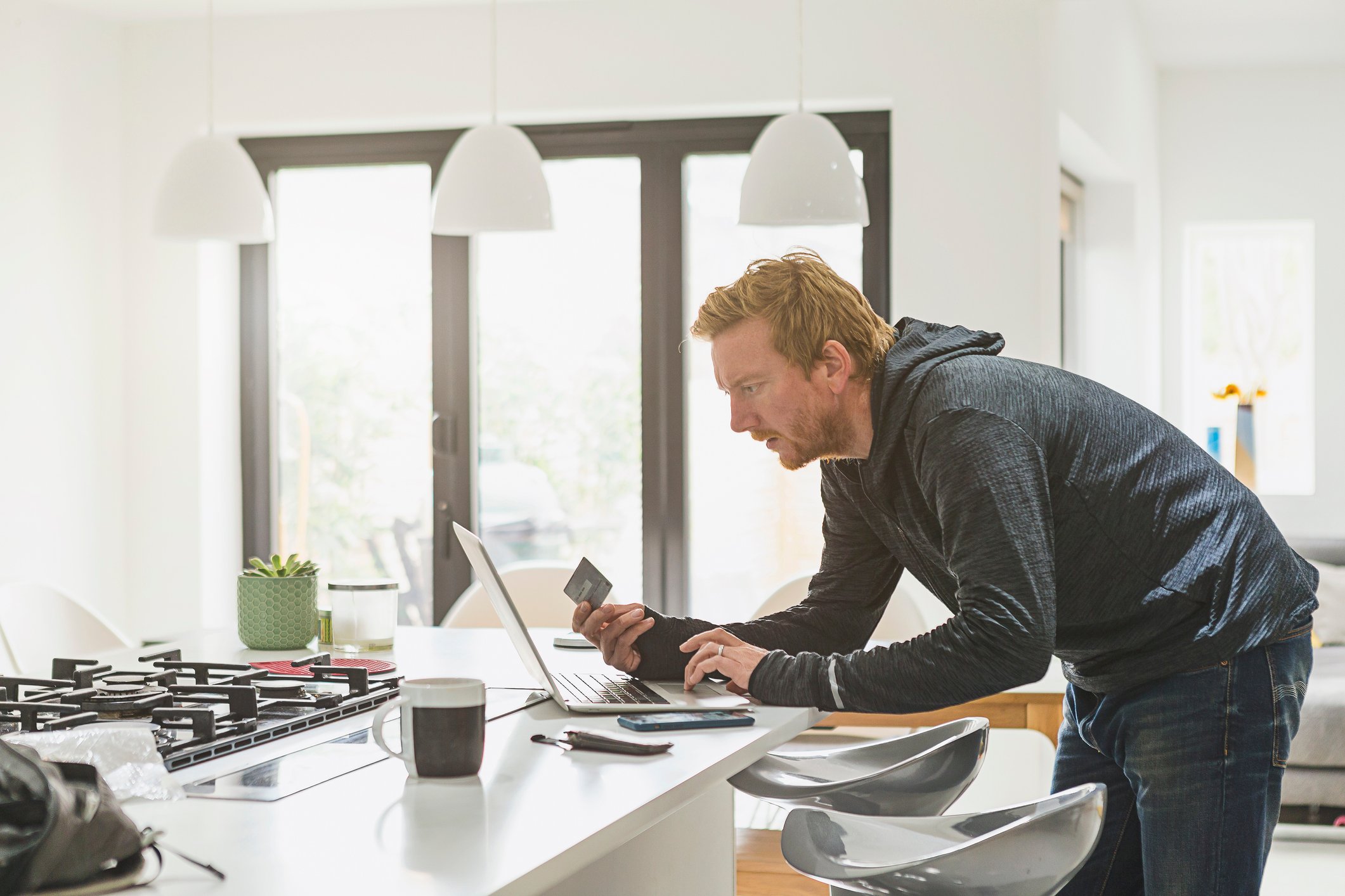 Man leaning over a laptop on a table while holding a credit card in his right hand.