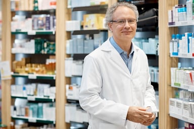 male pharmacist stands in front of shelf of drugs