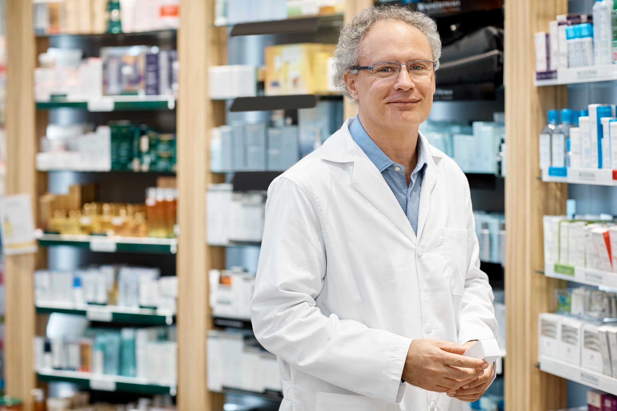 Male pharmacist stands in front of a large shelf of medications.