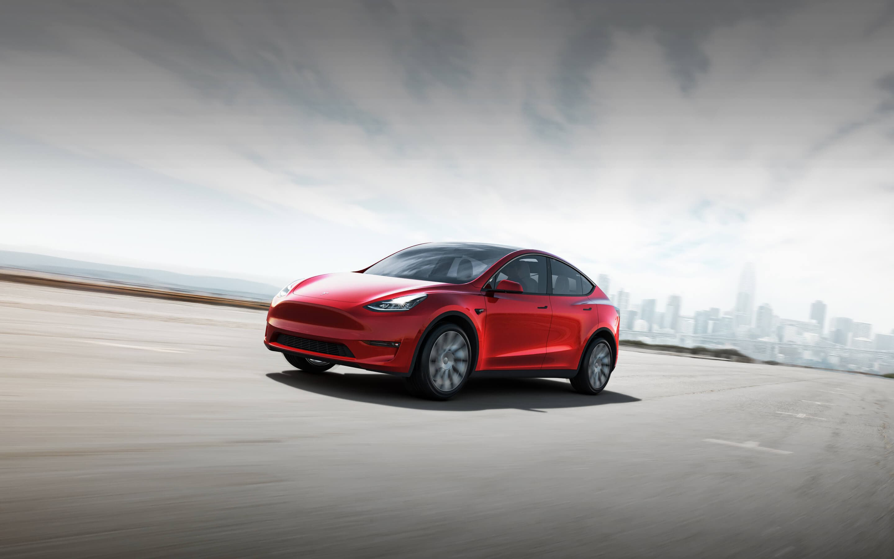 Red Tesla Model Y SUV on concrete pavement with a city skyline behind.