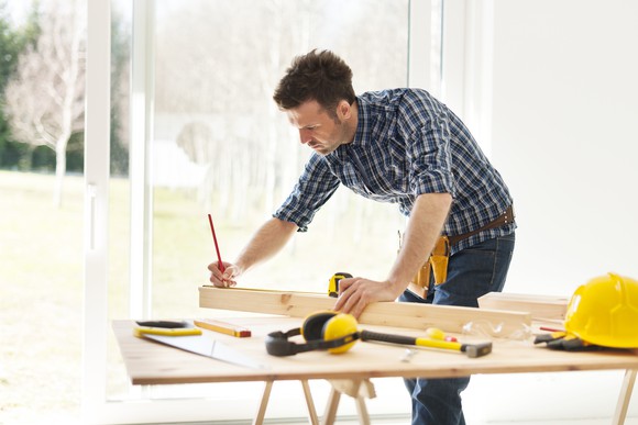 A man marks a measurement on a piece of wood on a work bench.