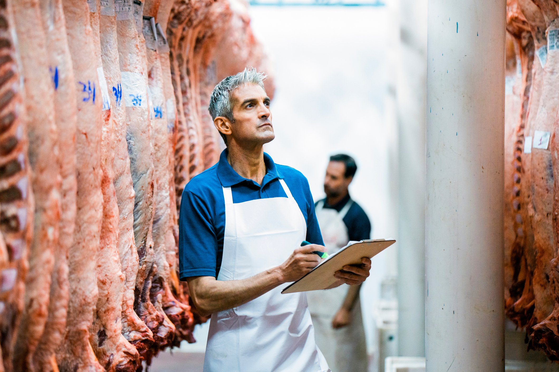 Inspectors in white aprons examine hanging sides of beef in a meatpacking plant.