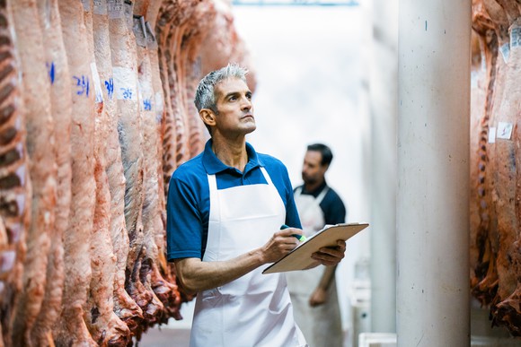 Inspectors in white aprons examine hanging sides of beef in a meatpacking plant.