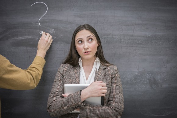 A woman in front of a blackboard holding a notebook, looking off to the side, while someone writes a question mark on the blackboard.