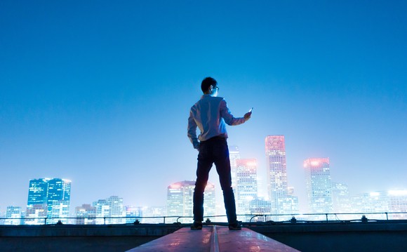 A young man looks at his phone before a night cityscape of a Chinese city. 