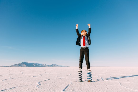 A young boy in a business suit stands on springs in the middle of a desert.