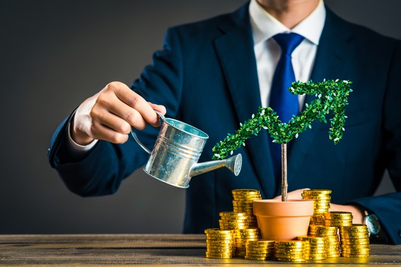 A man watering a plant with stacks of gold coins next to it. 
