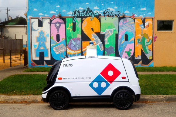 A Domiino's-branded Nuro R2 delivery robot parked at the curb in front of a colorful Houston sign.
