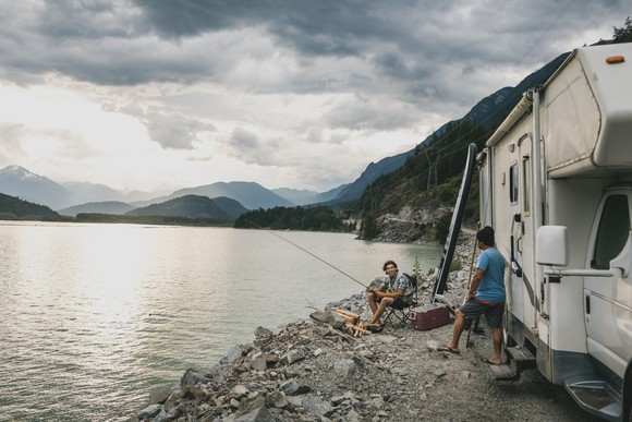 A family fishing with their RV parked on a lakeshore, with a cloudy, mountainous background.