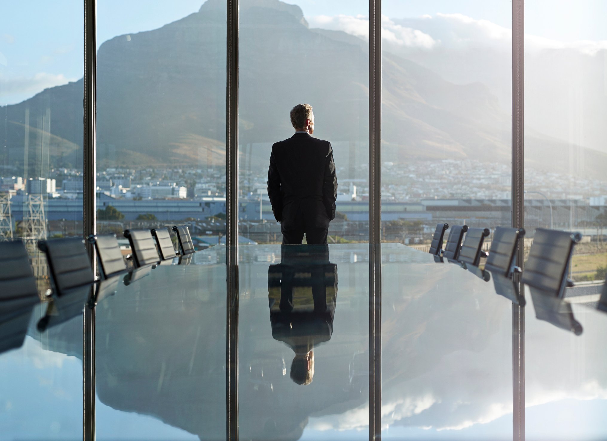 An executive looking out pensively from high windows over a cityscape to a distant mountain ridge.