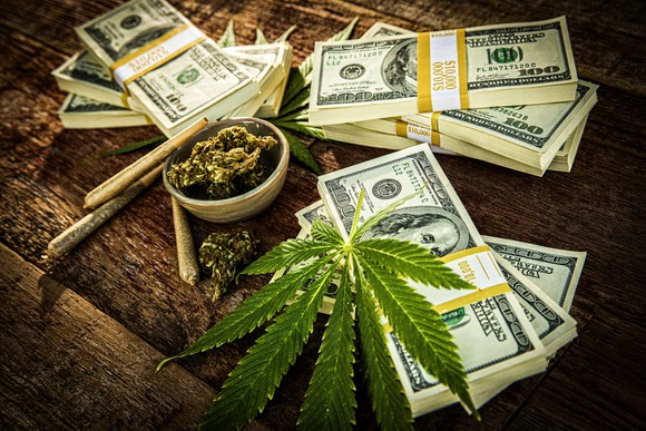 Piles of cash among cannabis buds and joints on a table.