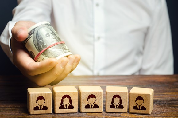 A man holds a roll of cash over blocks representing employees.