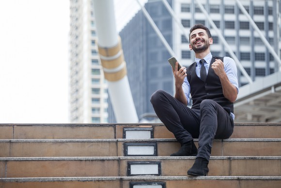 A man sitting on an outdoor stairway smiles and pumps his fist as he holds a cell phone.