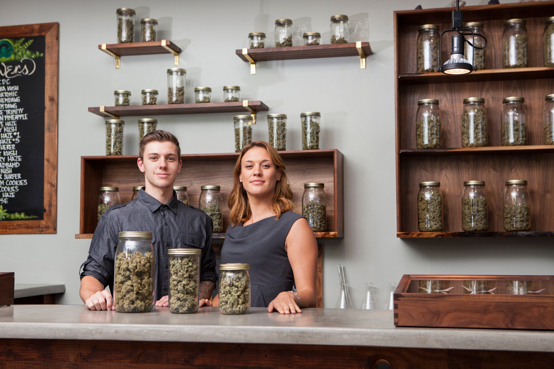 A pair of shopkeepers at a cannabis dispensary stand triumphantly in front of three jars filled with cannabis flower.