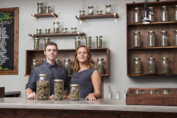 A pair of shopkeepers at a cannabis dispensary stand triumphantly in front of three jars filled with cannabis flower.