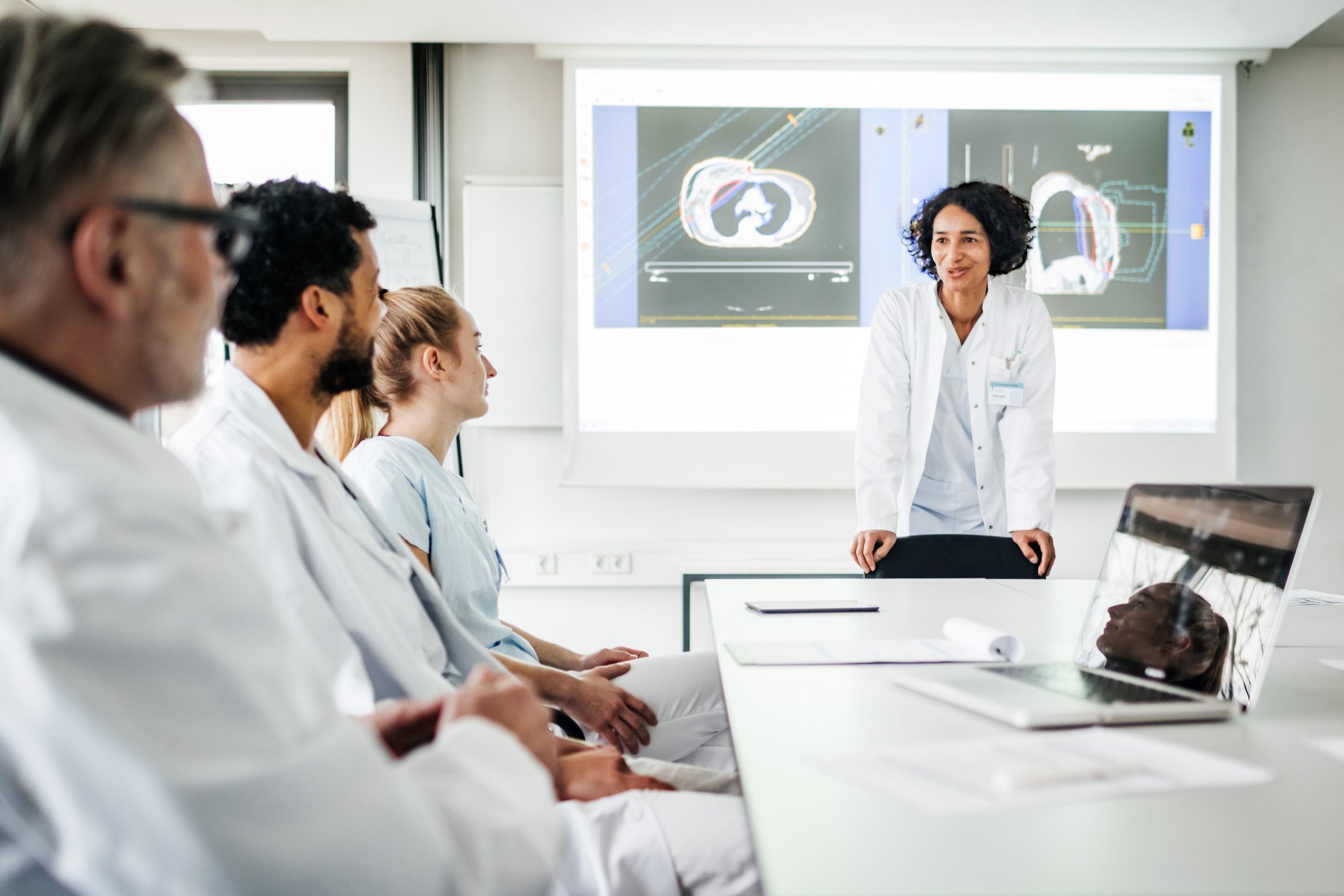 Two female and two male doctors in white coats sit around a table discussing data and medical scans.
