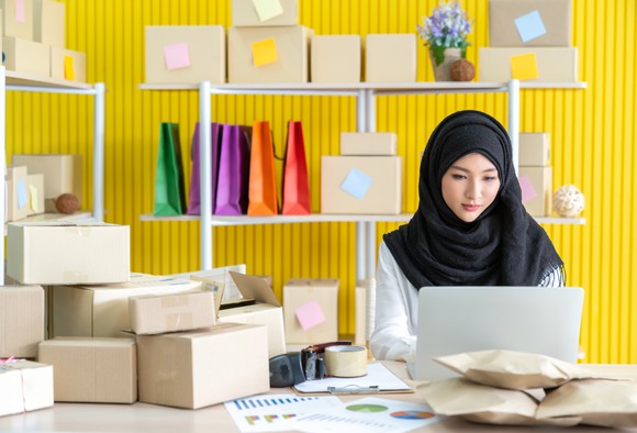 A woman in a headscarf uses a laptop at her small business to prepare to ship boxes of goods.