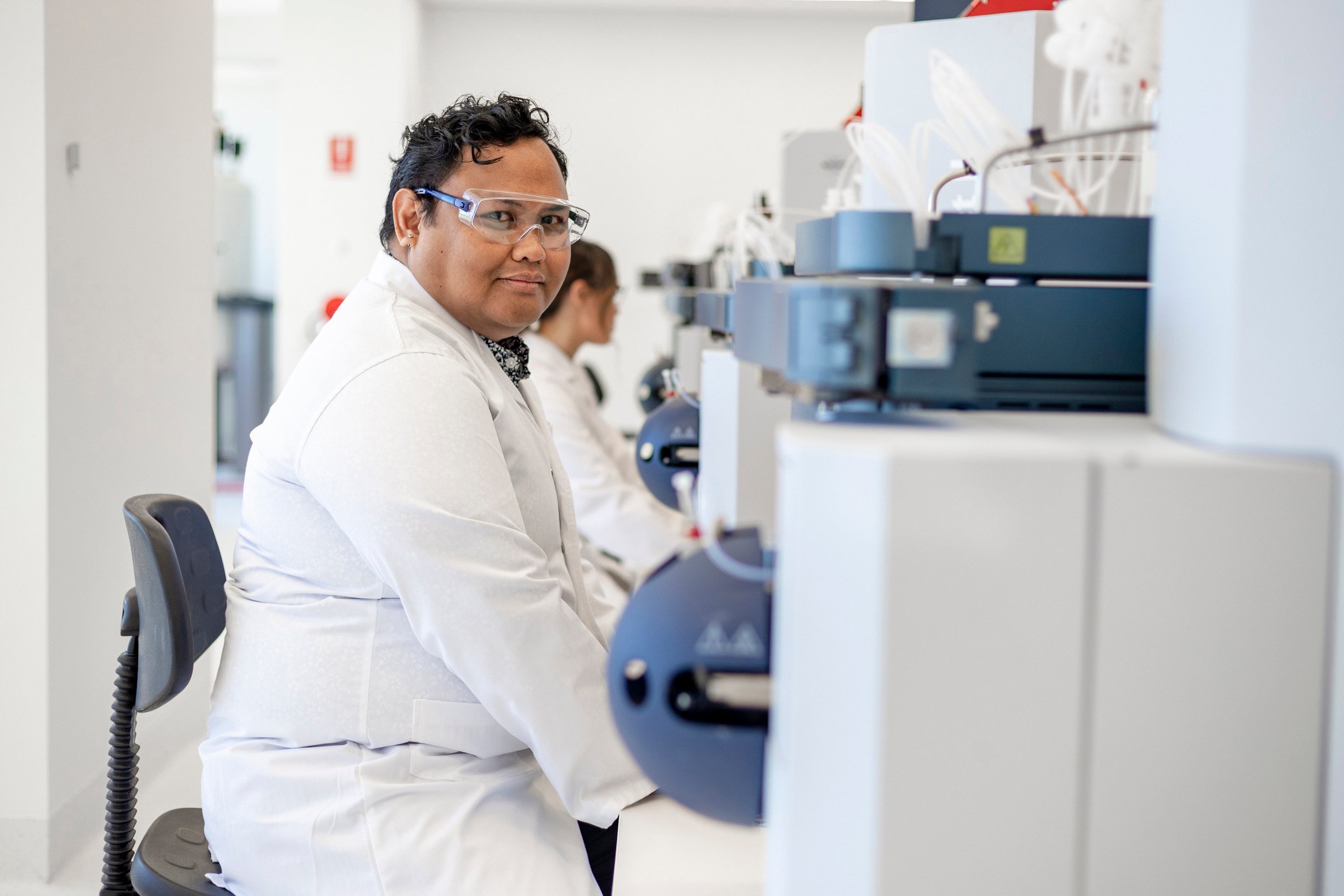 Male research in white coat and goggles sits at a desk in front of machinery in a laboratory setting.