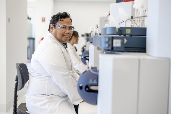 Male research in white coat and goggles sits at a desk in front of machinery in a laboratory setting.
