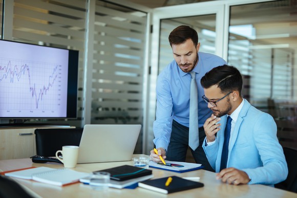 Two men in shirts and ties work on analysis at a desk with a computer screen displaying a chart.