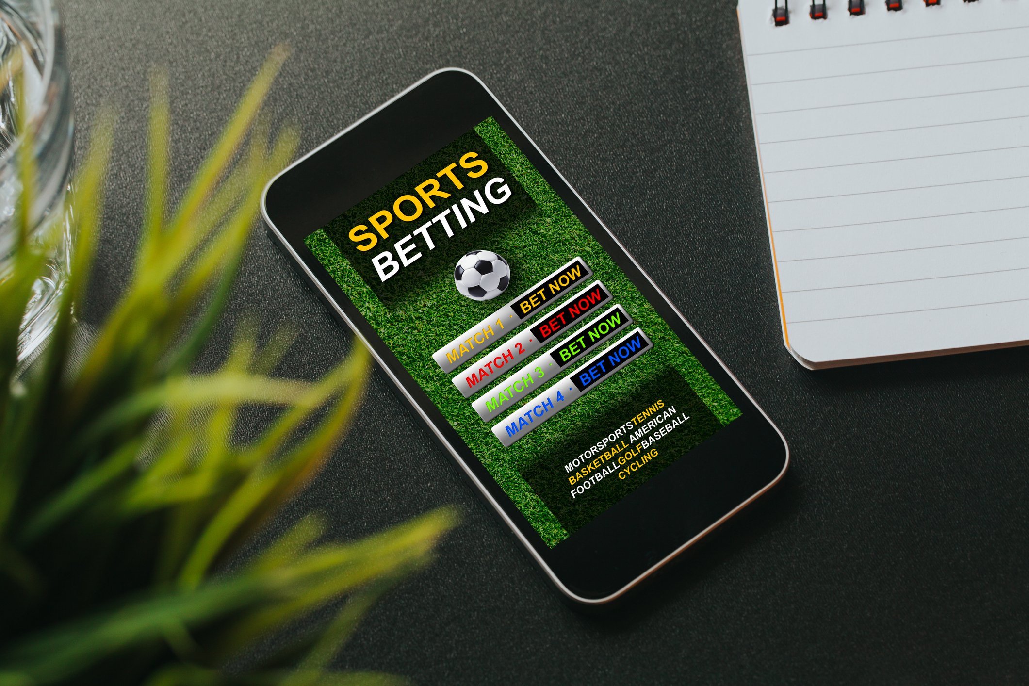 A smartphone with a sports betting app on its screen resting on a desktop between a notepad and a potted plant.