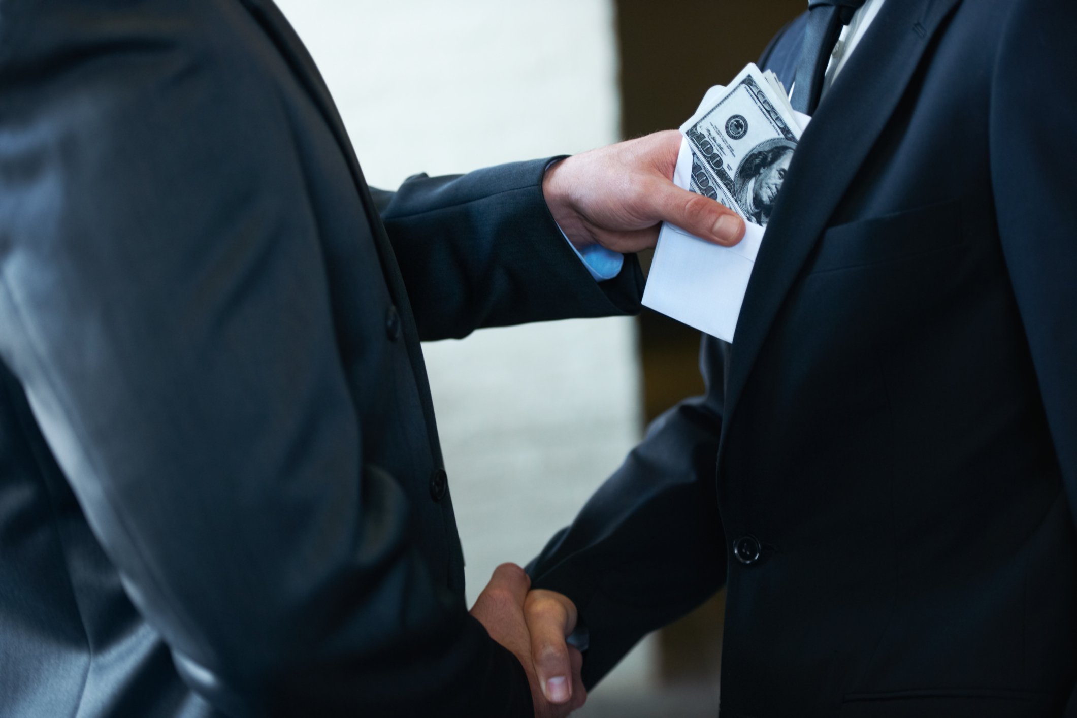 Two businessmen shaking hands with one stuffing an envelope of money into the front of the other's suit.
