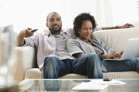 A couple sitting on a couch. He has a remote in his hand as he looks at a TV, and she's looking at a laptop computer.
