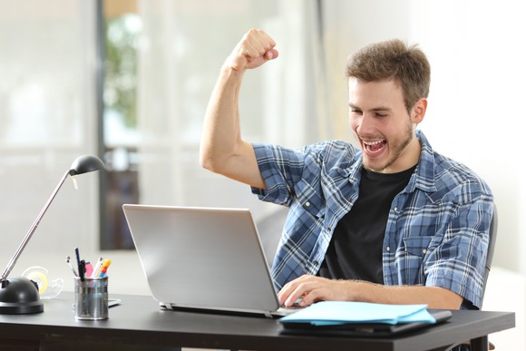 A man looks at his laptop and pumps his fist in celebration.